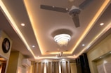 A modern ceiling design with recessed lighting and a central chandelier featuring hanging crystals. There are two ceiling fans with metallic blades beside the chandelier. The walls are adorned with a decorative clock and an air conditioning unit mounted above curtains.