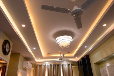 A modern ceiling design with recessed lighting and a central chandelier featuring hanging crystals. There are two ceiling fans with metallic blades beside the chandelier. The walls are adorned with a decorative clock and an air conditioning unit mounted above curtains.
