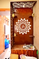 A wooden corner with intricate lattice patterns and a central design resembling a floral shape with the Om symbol. It features decorative hanging lamps and shelves with golden accent pieces. Clutter, including a cardboard box and scattered items, is visible on the floor and surrounding area.