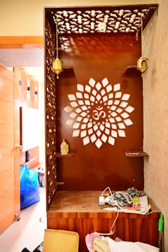 A wooden corner with intricate lattice patterns and a central design resembling a floral shape with the Om symbol. It features decorative hanging lamps and shelves with golden accent pieces. Clutter, including a cardboard box and scattered items, is visible on the floor and surrounding area.