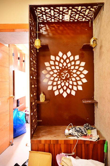 A wooden corner with intricate lattice patterns and a central design resembling a floral shape with the Om symbol. It features decorative hanging lamps and shelves with golden accent pieces. Clutter, including a cardboard box and scattered items, is visible on the floor and surrounding area.