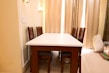 A dining area featuring a rectangular white table surrounded by six wooden chairs with high backs. The room is softly lit by wall lamps and natural light filtered through sheer curtains. The walls are cream-colored with decorative paneling. Bags and miscellaneous items are placed in the corner.