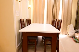 A dining area featuring a rectangular white table surrounded by six wooden chairs with high backs. The room is softly lit by wall lamps and natural light filtered through sheer curtains. The walls are cream-colored with decorative paneling. Bags and miscellaneous items are placed in the corner.