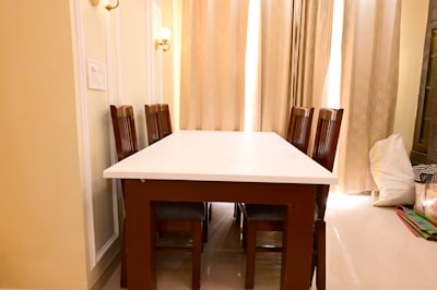 A dining area featuring a rectangular white table surrounded by six wooden chairs with high backs. The room is softly lit by wall lamps and natural light filtered through sheer curtains. The walls are cream-colored with decorative paneling. Bags and miscellaneous items are placed in the corner.
