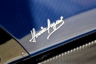 A close-up view of a car's surface showcasing a shiny metallic signature engraved on a sleek blue and carbon fiber material. The script style of the signature is elegant and prominently displayed.