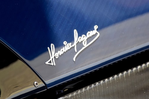 A close-up view of a car's surface showcasing a shiny metallic signature engraved on a sleek blue and carbon fiber material. The script style of the signature is elegant and prominently displayed.