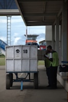 Two workers are standing next to a large white cart labeled with the number 10. They appear to be checking or loading items. The scene is set outdoors near an airport, with a terminal building and control tower visible in the background.
