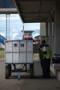 Two workers are standing next to a large white cart labeled with the number 10. They appear to be checking or loading items. The scene is set outdoors near an airport, with a terminal building and control tower visible in the background.