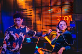 A woman in a dark dress is holding a phone receiver, looking to the side with an attentive expression. Beside her, a man in a colorful shirt rests his hand on a nearby arcade machine, looking directly at the camera. The room is dimly lit with vibrant orange and blue neon lights reflecting on the walls and window blinds.