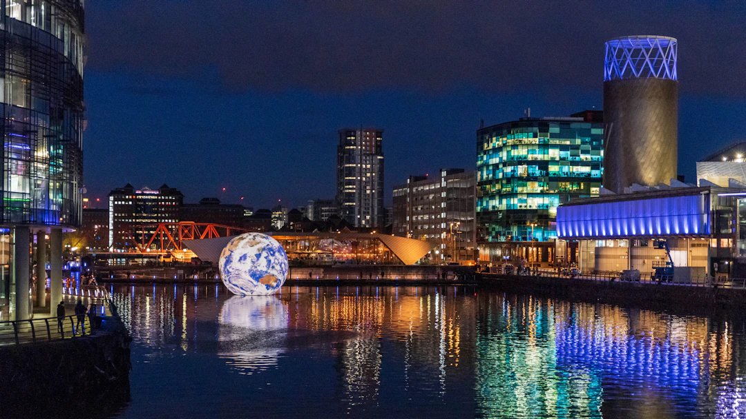 Waterfront buildings and a lit globe sculpture reflected in still water at night
