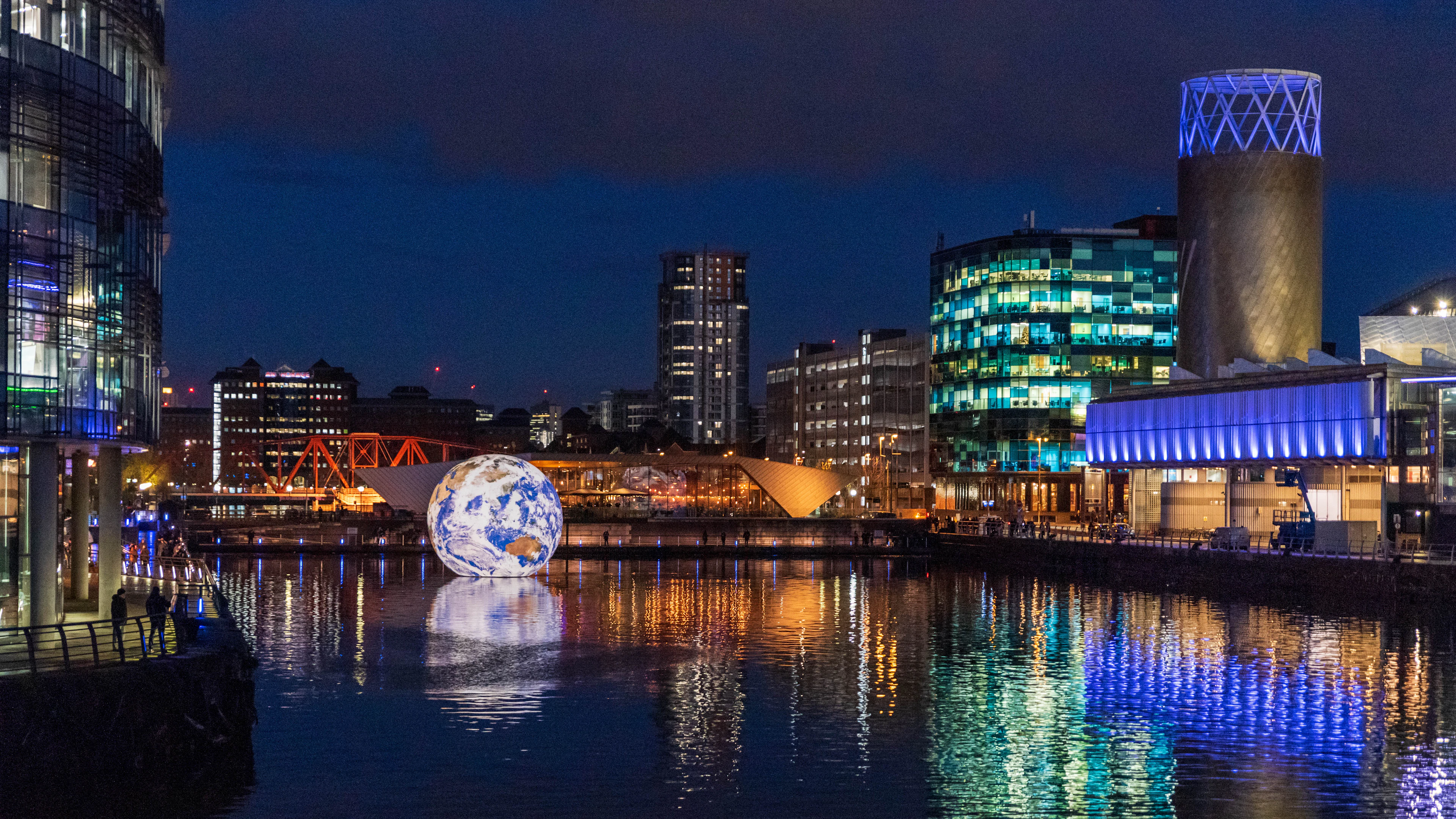 Salford Quays at night, the lit towers and glass frontages of MediaCityUK doubled in still black water beside a glowing floating globe.