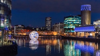 A nighttime cityscape featuring a large illuminated globe floating on a river. Surrounding this, various modern buildings with glass facades are lit with vibrant lights, reflecting on the water. The scene includes a distinctive tower with a blue-lit top and a bright red bridge in the background.