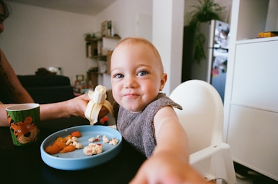 A toddler with blue eyes sits in a high chair, eating from a blue plate containing pieces of food and a sliced banana held by an adult's hand. The child appears cheerful and is reaching out towards the camera. A green cup with a tiger illustration is on the table. The background shows a home setting with a white wall, a refrigerator, and shelves with various items.
