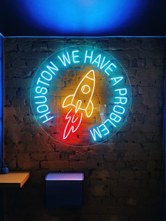 A neon sign on a brick wall featuring a rocket design and the text 'HOUSTON WE HAVE A PROBLEM' in bright, glowing colors.