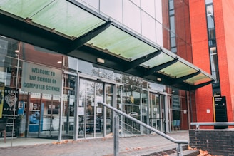 The entrance to a modern building with glass doors and a glass canopy. A sign reads 'Welcome to the School of Computer Science'. The building features red brick and glass with reflections of surrounding trees. A handrail leads up to the doors, and there are autumn leaves scattered on the pavement.
