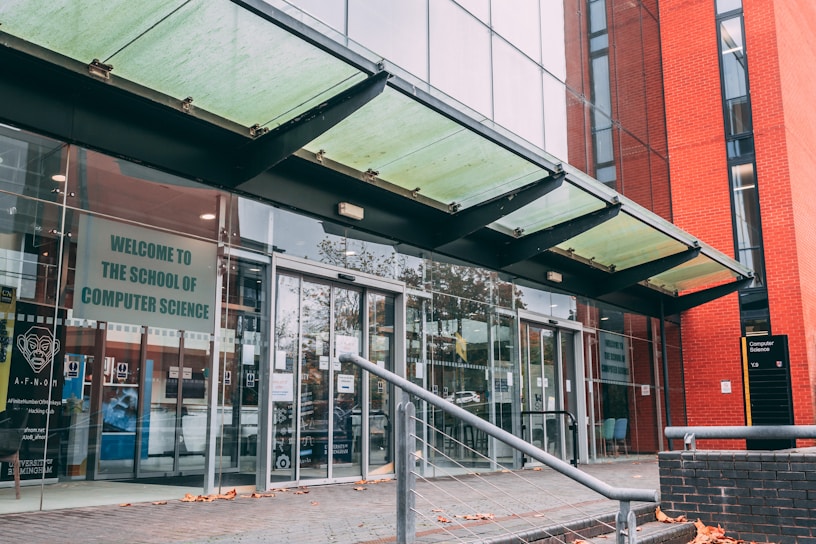 The entrance to a modern building with glass doors and a glass canopy. A sign reads 'Welcome to the School of Computer Science'. The building features red brick and glass with reflections of surrounding trees. A handrail leads up to the doors, and there are autumn leaves scattered on the pavement.