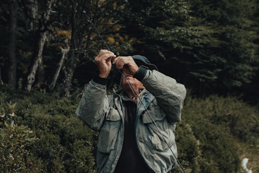 A person is adjusting a fishing line against a backdrop of dense, leafy greenery. Dressed in a light gray jacket and a dark hat, their face is mostly obscured. The setting feels like a forested area, conveying a sense of solitude and focus.