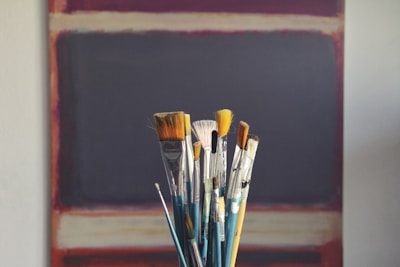 A collection of paintbrushes with various bristle shapes and sizes is arranged in a container, set against a blurred abstract painting in the background with dark and muted colors.