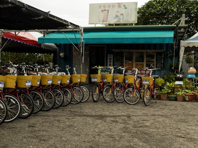 A row of rental bicycles with yellow baskets is neatly lined up in front of a small shop with a teal awning. The shop is surrounded by potted plants and features a sign with text in a foreign language. The setting suggests an outdoor recreational activity area.