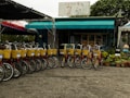 A row of rental bicycles with yellow baskets is neatly lined up in front of a small shop with a teal awning. The shop is surrounded by potted plants and features a sign with text in a foreign language. The setting suggests an outdoor recreational activity area.