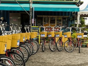 A row of bicycles with yellow baskets is parked in front of a small storefront. The storefront has a blue and green overhang and displays various signs in what appears to be Asian script. The area is surrounded by plants, and there are signs with numbers attached to each bicycle basket.