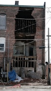 A brick building has suffered significant structural damage, with a large section of the wall missing, exposing its interior. The area around the building is cluttered with debris, including bricks and various wooden boards. Electrical wires are hanging across the scene.