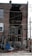 A brick building has suffered significant structural damage, with a large section of the wall missing, exposing its interior. The area around the building is cluttered with debris, including bricks and various wooden boards. Electrical wires are hanging across the scene.