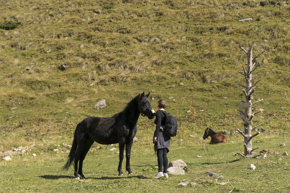 a man standing next to a black horse on a lush green hillside