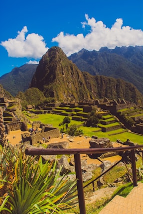 A breathtaking view of ancient stone structures set against a backdrop of lush green terraces. Majestic mountains rise dramatically in the distance under a clear, vibrant blue sky with fluffy clouds. The foreground features green plants and a wooden railing.