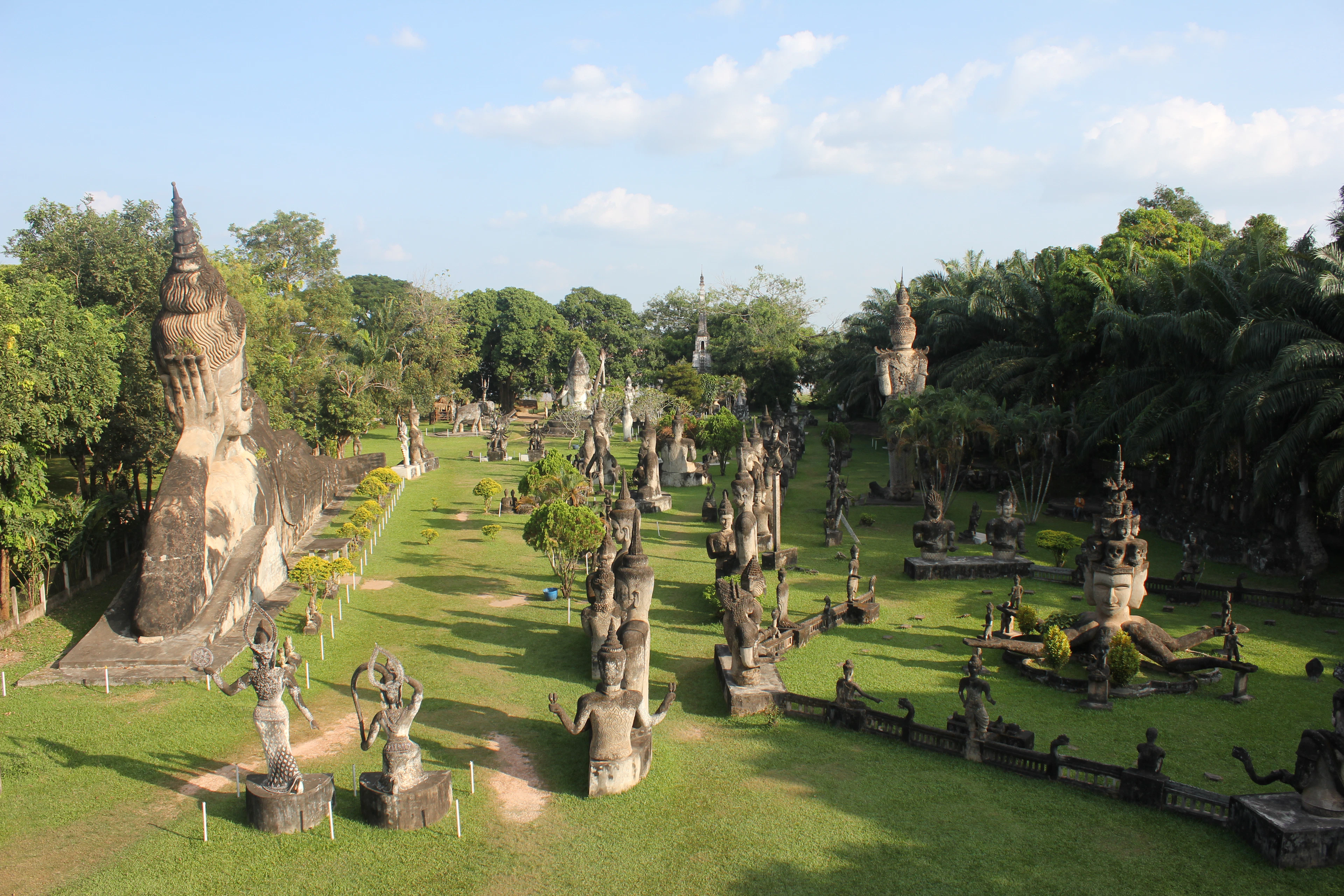 The sculpture meadow of Buddha Park outside Vientiane, dozens of weathered concrete Buddhas and Hindu deities standing across a sunlit green lawn framed by palms