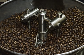 A large quantity of roasted coffee beans in a circular roasting machine. The machine features metallic components, including stirring arms and stabilizing structures, designed to agitate and evenly roast the beans.