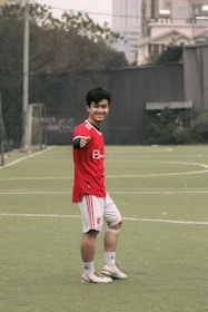A person wearing a red sports jersey and white shorts is standing on a grassy sports field, posing and smiling towards the camera. They are making a gesture with their left hand. In the background, there are buildings, trees, and a goal post visible, suggesting an outdoor urban environment.