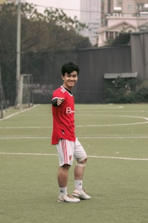 A person wearing a red sports jersey and white shorts is standing on a grassy sports field, posing and smiling towards the camera. They are making a gesture with their left hand. In the background, there are buildings, trees, and a goal post visible, suggesting an outdoor urban environment.