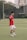 A person wearing a red sports jersey and white shorts is standing on a grassy sports field, posing and smiling towards the camera. They are making a gesture with their left hand. In the background, there are buildings, trees, and a goal post visible, suggesting an outdoor urban environment.