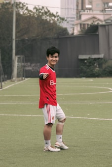 A person wearing a red sports jersey and white shorts is standing on a grassy sports field, posing and smiling towards the camera. They are making a gesture with their left hand. In the background, there are buildings, trees, and a goal post visible, suggesting an outdoor urban environment.