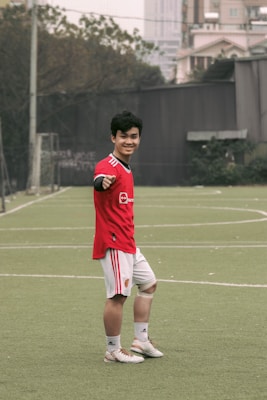 A person wearing a red sports jersey and white shorts is standing on a grassy sports field, posing and smiling towards the camera. They are making a gesture with their left hand. In the background, there are buildings, trees, and a goal post visible, suggesting an outdoor urban environment.