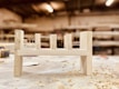 A partially assembled wooden furniture piece rests on a workshop table, surrounded by wood shavings. The background is a blurred view of a workshop, with shelves and tools visible.