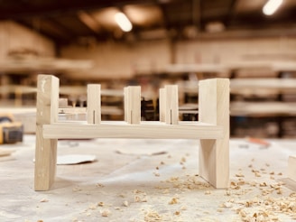 A partially assembled wooden furniture piece rests on a workshop table, surrounded by wood shavings. The background is a blurred view of a workshop, with shelves and tools visible.