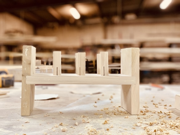 A partially assembled wooden furniture piece rests on a workshop table, surrounded by wood shavings. The background is a blurred view of a workshop, with shelves and tools visible.