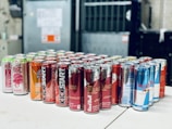 A collection of energy drink cans arranged on a white surface. The cans are from various brands, including Red Bull, Mountain Dew Kickstart, and Rise. They are organized in several rows, featuring a variety of colors and designs. The background is a blurred industrial setting.