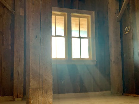 Sunlight streams through a window in a rustic wooden room, casting soft beams across the floor. The room is empty, with visible wooden beams and walls, creating a warm and serene atmosphere.