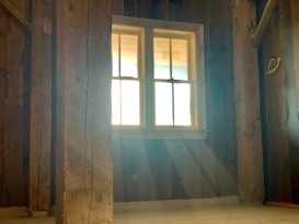 Sunlight streams through a window in a rustic wooden room, casting soft beams across the floor. The room is empty, with visible wooden beams and walls, creating a warm and serene atmosphere.