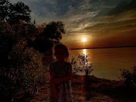 A silhouetted child stands by a tranquil lakeside during a sunset. The sky is painted with shades of orange and yellow, and the sun is near the horizon, reflecting off the water's surface. Trees and foliage frame the scene.