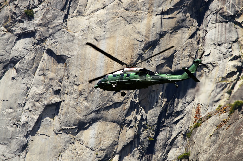 a green helicopter flying over a mountain side