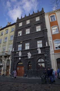 A black, ornate building with decorative architectural elements and multiple windows. Statues and reliefs adorn the facade above a wooden entrance with a sign in Cyrillic. Adjacent buildings differ in color and style. People are gathered in front, wearing winter clothing, suggesting a cold day.