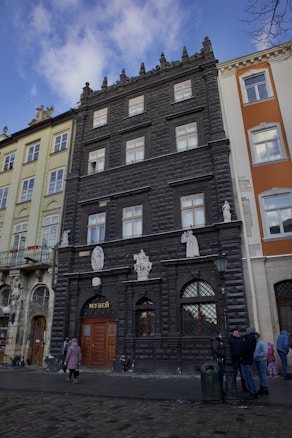 A black, ornate building with decorative architectural elements and multiple windows. Statues and reliefs adorn the facade above a wooden entrance with a sign in Cyrillic. Adjacent buildings differ in color and style. People are gathered in front, wearing winter clothing, suggesting a cold day.