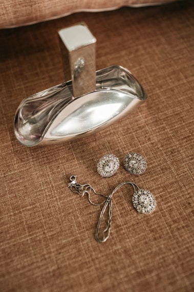 A silver-colored perfume bottle with a reflective surface is placed on a textured brown surface. Beside it, there is a pair of ornate, circular earrings with intricate detailing, as well as a matching pendant with a chain.