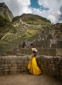 A young woman wearing a flowing turquoise dress with coral accessories, standing against a backdrop of ancient Greek ruins.