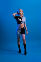 A person with long blonde hair poses confidently against a solid blue background. They are wearing a two-toned outfit consisting of a cropped top and shorts, featuring the word 'FLEX' across the top. The color scheme is predominantly black and light blue, complemented by black knee-high socks.