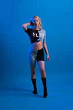 A person with long blonde hair poses confidently against a solid blue background. They are wearing a two-toned outfit consisting of a cropped top and shorts, featuring the word 'FLEX' across the top. The color scheme is predominantly black and light blue, complemented by black knee-high socks.